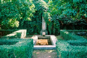 Une fontaine ancienne avec une statue au centre, entourée de haies vertes et d'une végétation luxuriante.